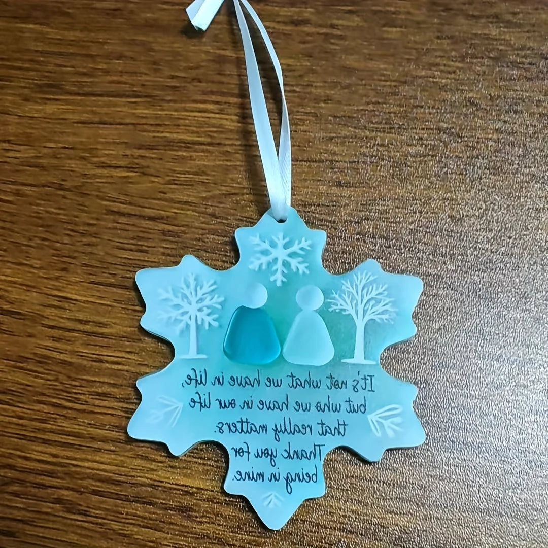 A light blue snowflake-shaped sea glass ornament with a white ribbon, featuring a silhouette of a person and a tree.