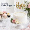 A white tiered cake decorated with floral accents and gold topper sticks on a white platter.