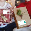 A person is holding a brown gift box tied with twine and a Christmas tag, with another red gift box and tags visible in the b