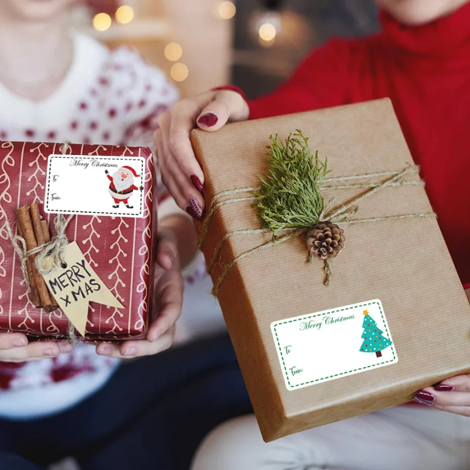 A person is holding a brown gift box tied with twine and a Christmas tag, with another red gift box and tags visible in the b