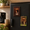 A dark refrigerator door features two framed wooden puzzle magnets depicting scenes for home decor.