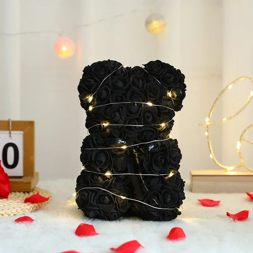 A black artificial rose teddy bear wrapped in silver wire, lying on white fluffy material with red petals.