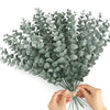A bouquet of artificial eucalyptus stems with silvery-green leaves being held by hands.