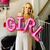 A smiling woman holds large pink foil letter balloons spelling 'GIRL' in a light-colored outfit.