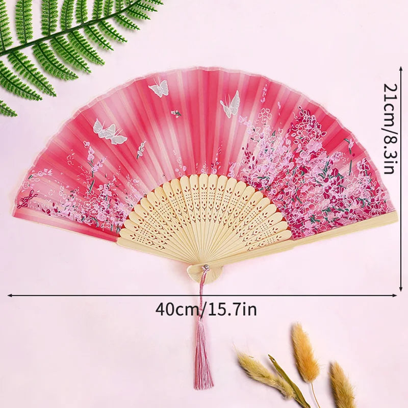 A pink and magenta folding fan with delicate floral patterns and a tassel, shown against a light pink background.