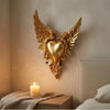 Gold ornate winged heart wall decor mounted on a beige wall above a wooden nightstand.