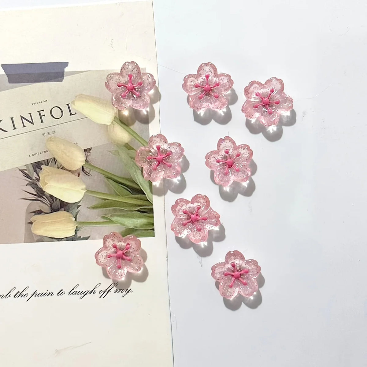 Pink cherry blossom fridge magnets scattered on a white background next to some dried foliage.