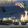 A set of black metal candle tins with lids placed on a wooden surface next to white candles and orchids.