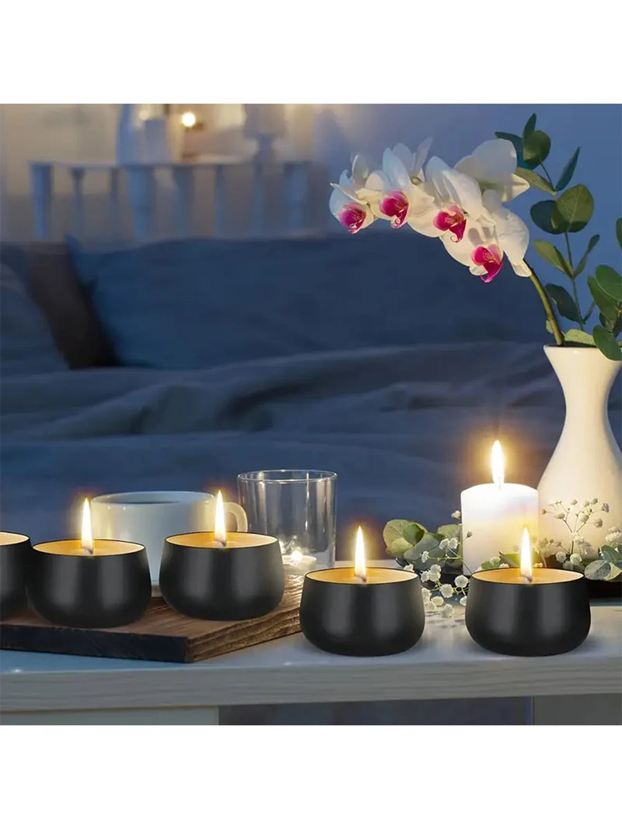 A set of black metal candle tins with lids placed on a wooden surface next to white candles and orchids.