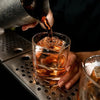 A hand pours amber liquid from a dark bottle into a glass filled with ice and whiskey.