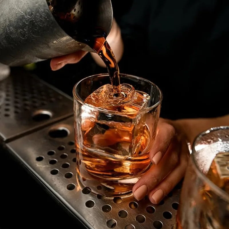 A hand pours amber liquid from a dark bottle into a glass filled with ice and whiskey.