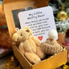 Un pequeño peluche de oso marrón claro con un corazón rojo y una tarjeta inspiradora dentro de una caja dorada.