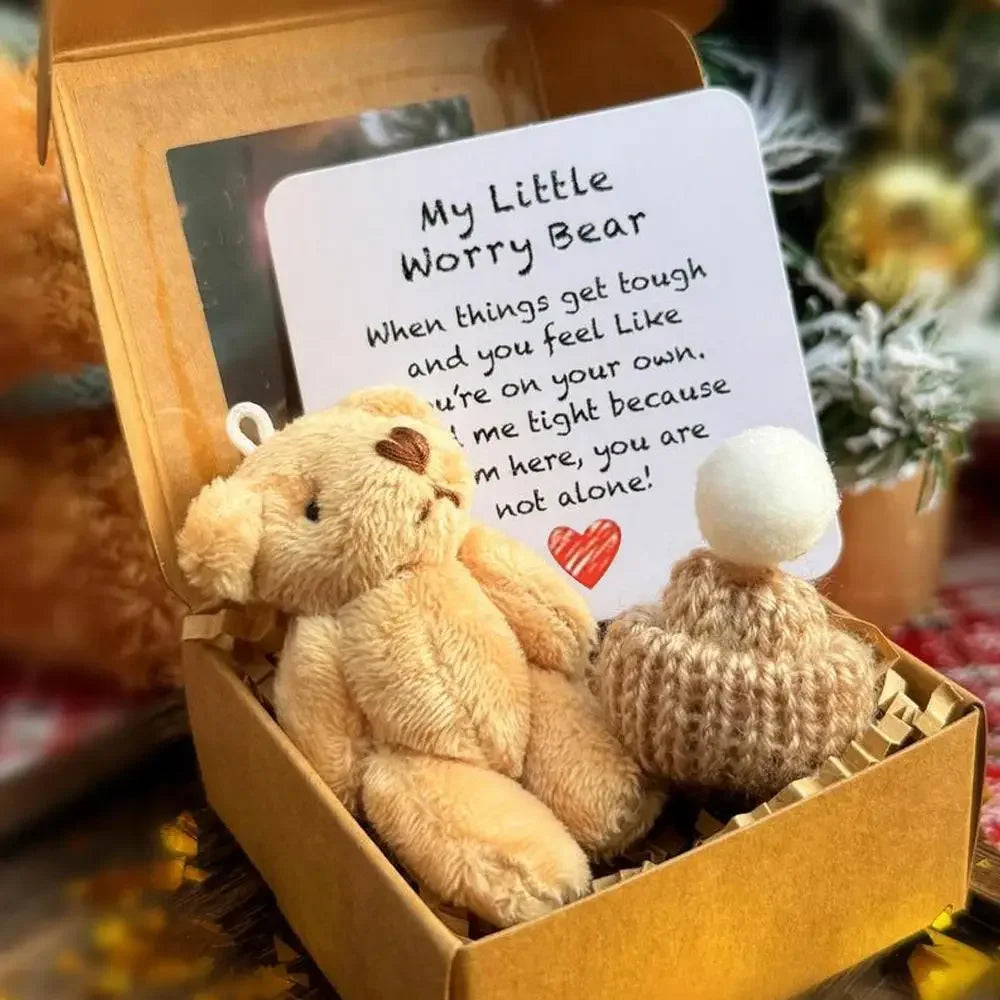 Un pequeño peluche de oso marrón claro con un corazón rojo y una tarjeta inspiradora dentro de una caja dorada.