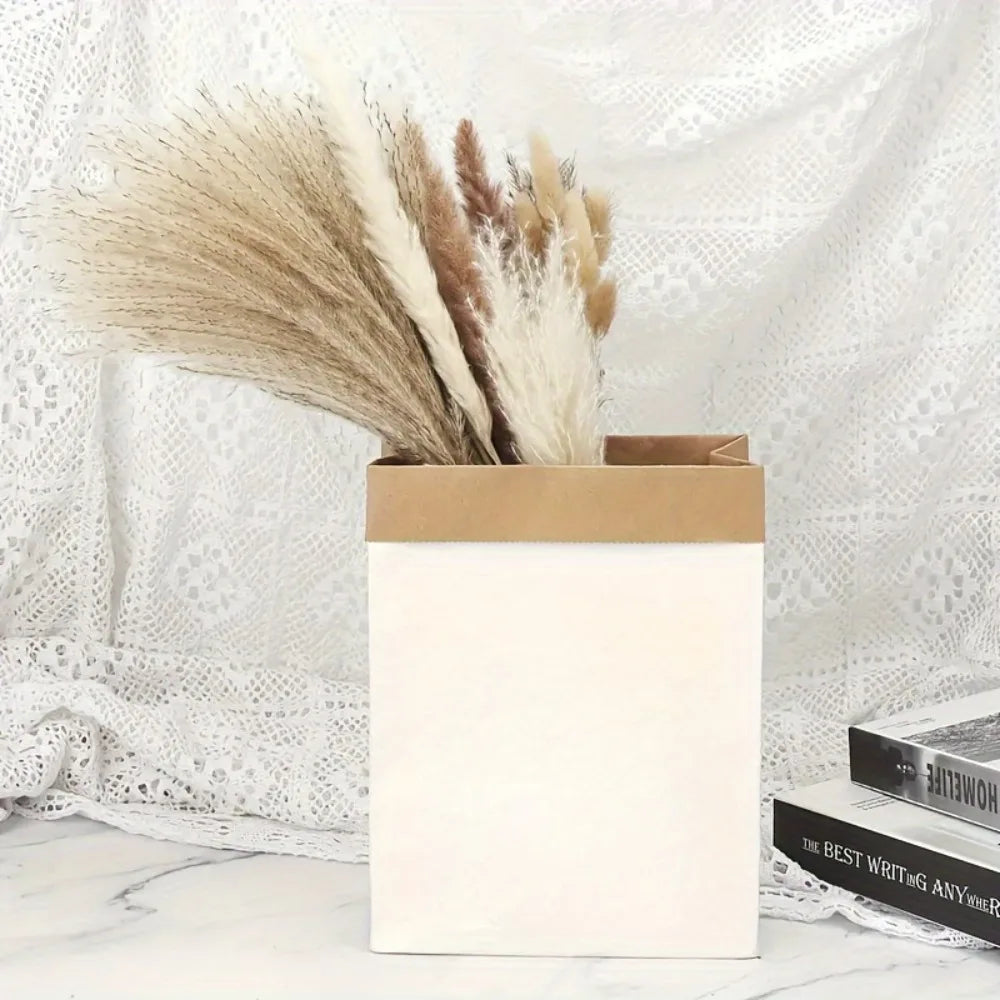 A bouquet of dried pampas grass flowers in various shades of beige and brown displayed in a white box against a sheer white b
