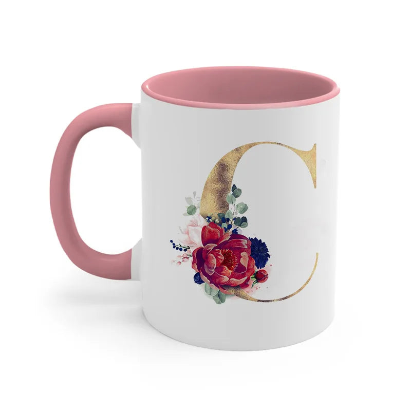 A pink ceramic coffee mug featuring a large gold letter C decorated with colorful floral accents.