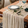 A beige cheesecloth table runner draped over a wooden table set with plates, silverware, and a glass of blue liquid.