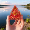 Hand holding a clear resin pyramid filled with red stones and a copper coil against a lake background