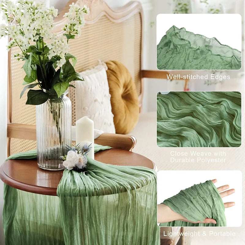Sage green gauze table runner draped over a wooden table with a floral arrangement in a glass vase.