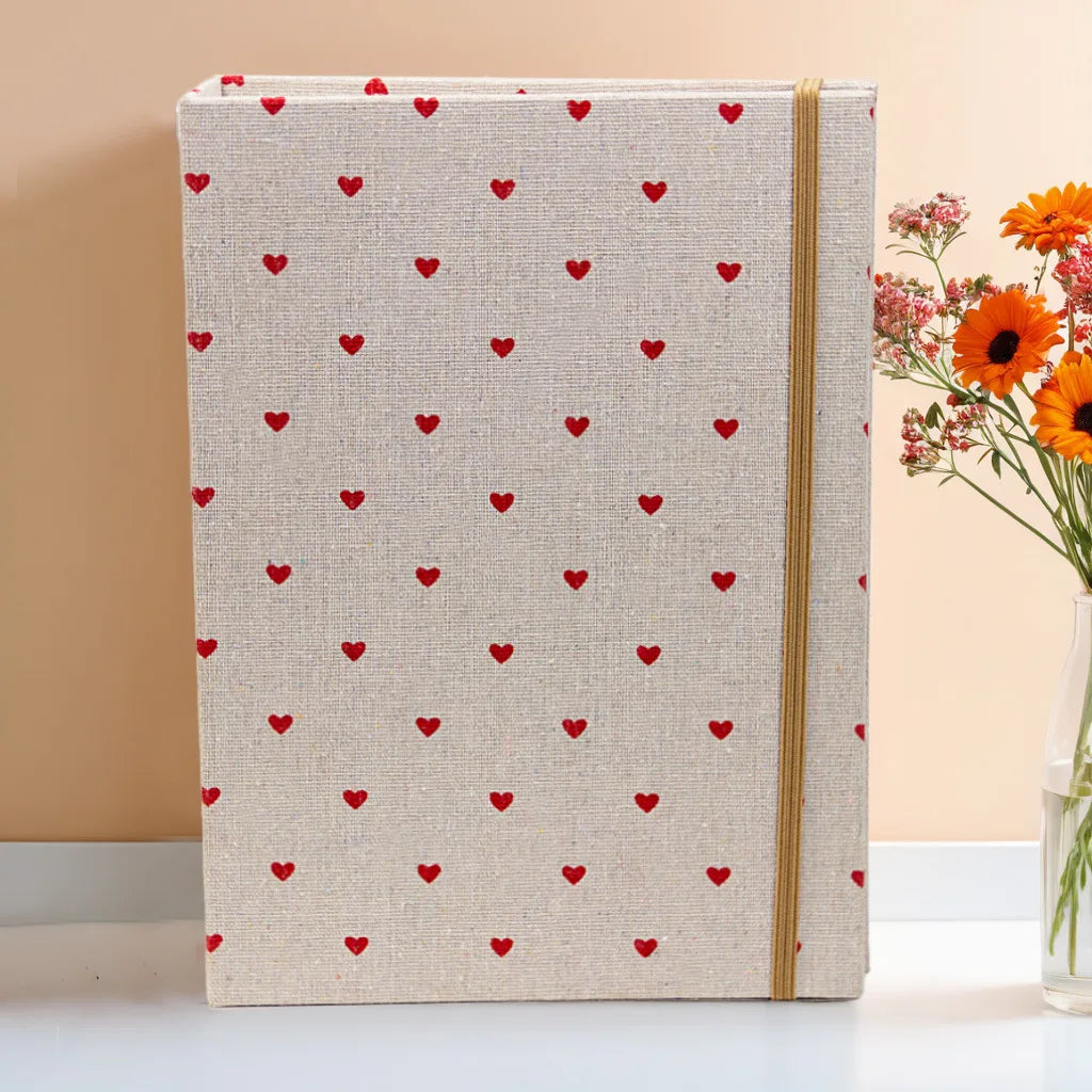 Beige Fotobuch mit roten Herzmustern und goldenem Einband auf weißer Fläche.