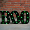 Letrero negro en forma de BOO con luces LED verdes brillantes contra una pared de ladrillos.