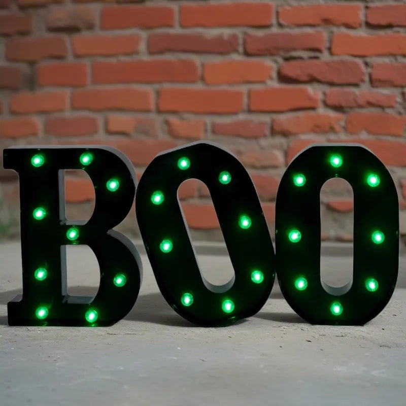 Letrero negro en forma de BOO con luces LED verdes brillantes contra una pared de ladrillos.