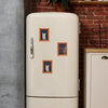A cream-colored fridge magnet featuring three framed illustrations of cats on a white cabinet.