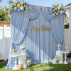 A blue satin backdrop curtain decorated with floral arrangements and gold balloons for a birthday party or photoshoot.