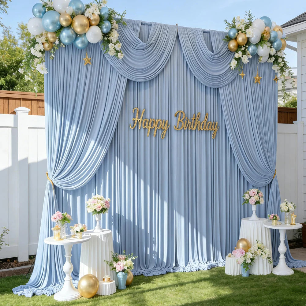 A blue satin backdrop curtain decorated with floral arrangements and gold balloons for a birthday party or photoshoot.