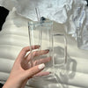 A hand holds a clear insulated glass coffee mug with a lid and straw on a white surface.