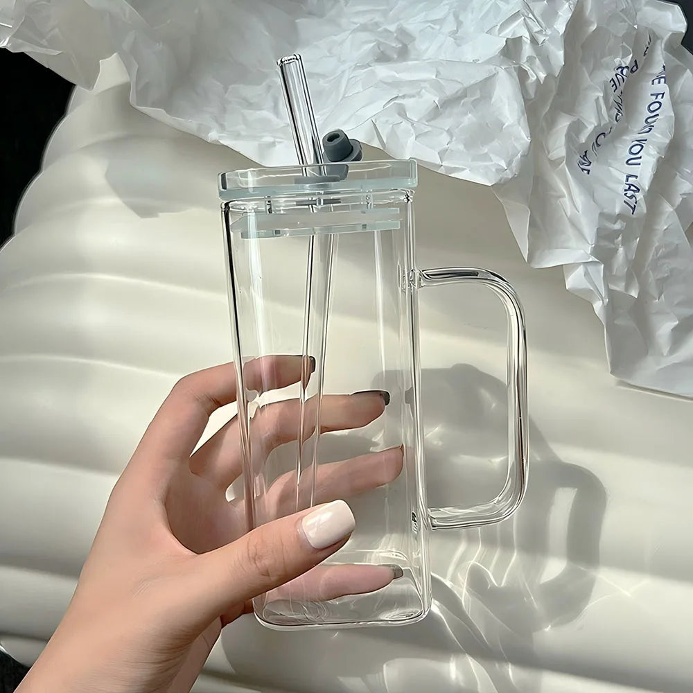 A hand holds a clear insulated glass coffee mug with a lid and straw on a white surface.