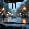 A silver cross pendant on a black beaded chain hanging in the rain inside a car.
