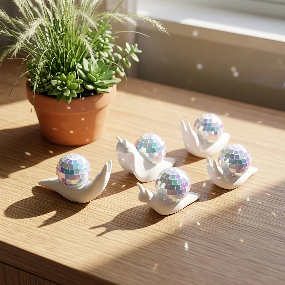 A white snail shaped sun catcher with a disco ball on the shell rests on a wooden surface next to a potted plant.