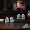 Three small gray stone monk statues with closed eyes sitting on a dark wooden surface.