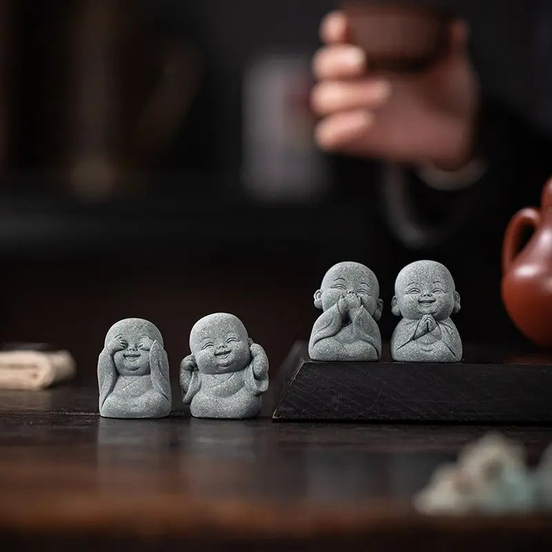 Three small gray stone monk statues with closed eyes sitting on a dark wooden surface.