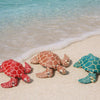 Three 3D printed articulated turtle figurines resting on a sandy beach near clear blue water.