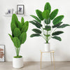 A large artificial Bird of Paradise plant with lush green leaves displayed in a white pot on a gold-toned stand.