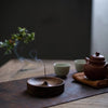 A dark wood incense stick is burning in a shallow wooden bowl on a brown slate coaster.