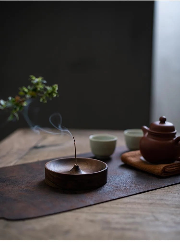 A dark wood incense stick is burning in a shallow wooden bowl on a brown slate coaster.
