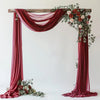 A draped archway setup featuring rich burgundy chiffon fabric and floral greenery for a wedding or event backdrop.
