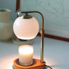 A brass aromatherapy wax melting lamp with a white spherical light illuminates a candle on a wooden base.