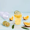 A glass of yellow citrus drink with lime slices and orange slices on a light wooden surface.