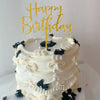 A white buttercream cake decorated with black floral accents and a gold Happy Birthday topper.