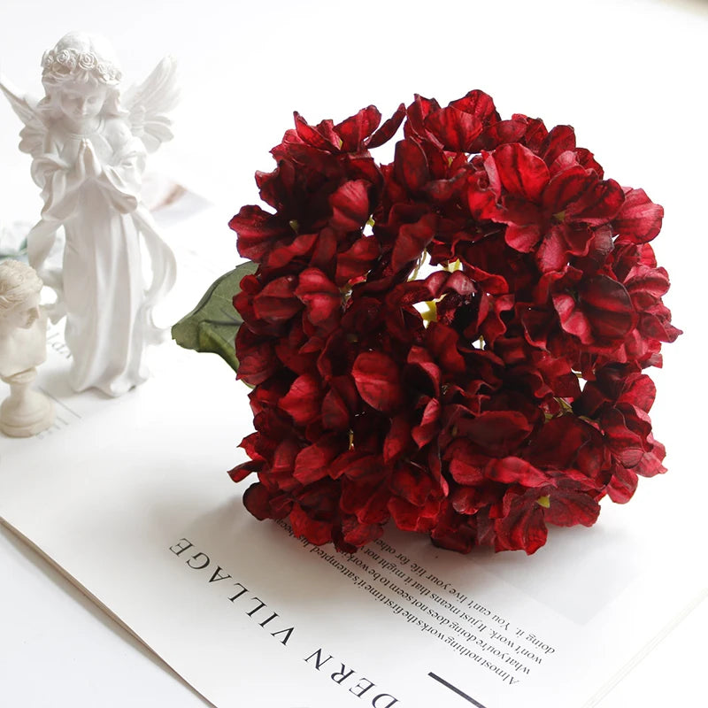 A bouquet of deep red silk hydrangea stems with vintage burnt edge petals displayed on a white surface.