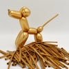 A golden latex modeling balloon shaped like a stylized animal standing on a pile of thin, gold-colored straws.
