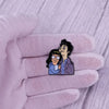A hand wearing a light pink glove holds an enamel pin badge featuring two cartoon characters.