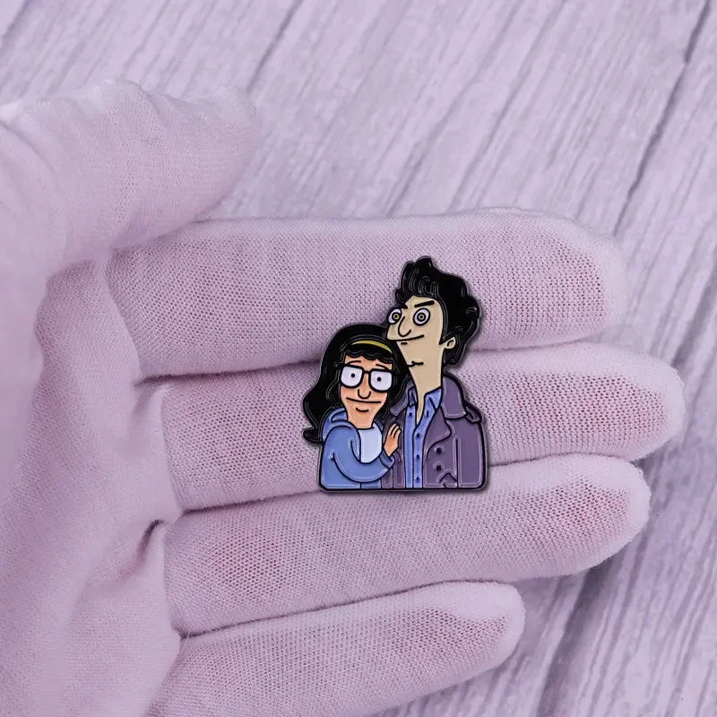A hand wearing a light pink glove holds an enamel pin badge featuring two cartoon characters.