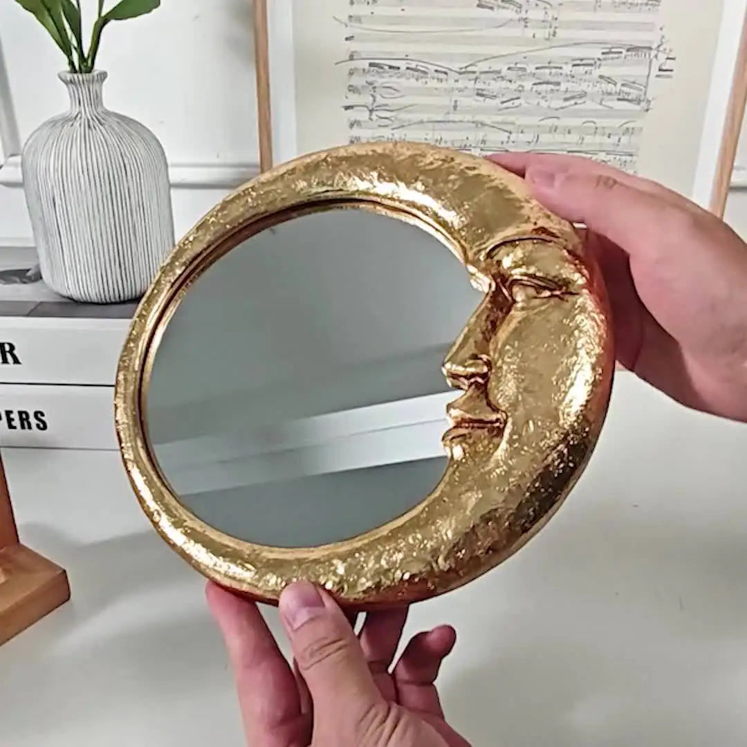 A hand holds a round, gold resin decorative hanging mirror shaped like a crescent moon.
