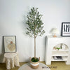 A tall artificial olive tree branches displayed against a white wall next to a small white shelf.