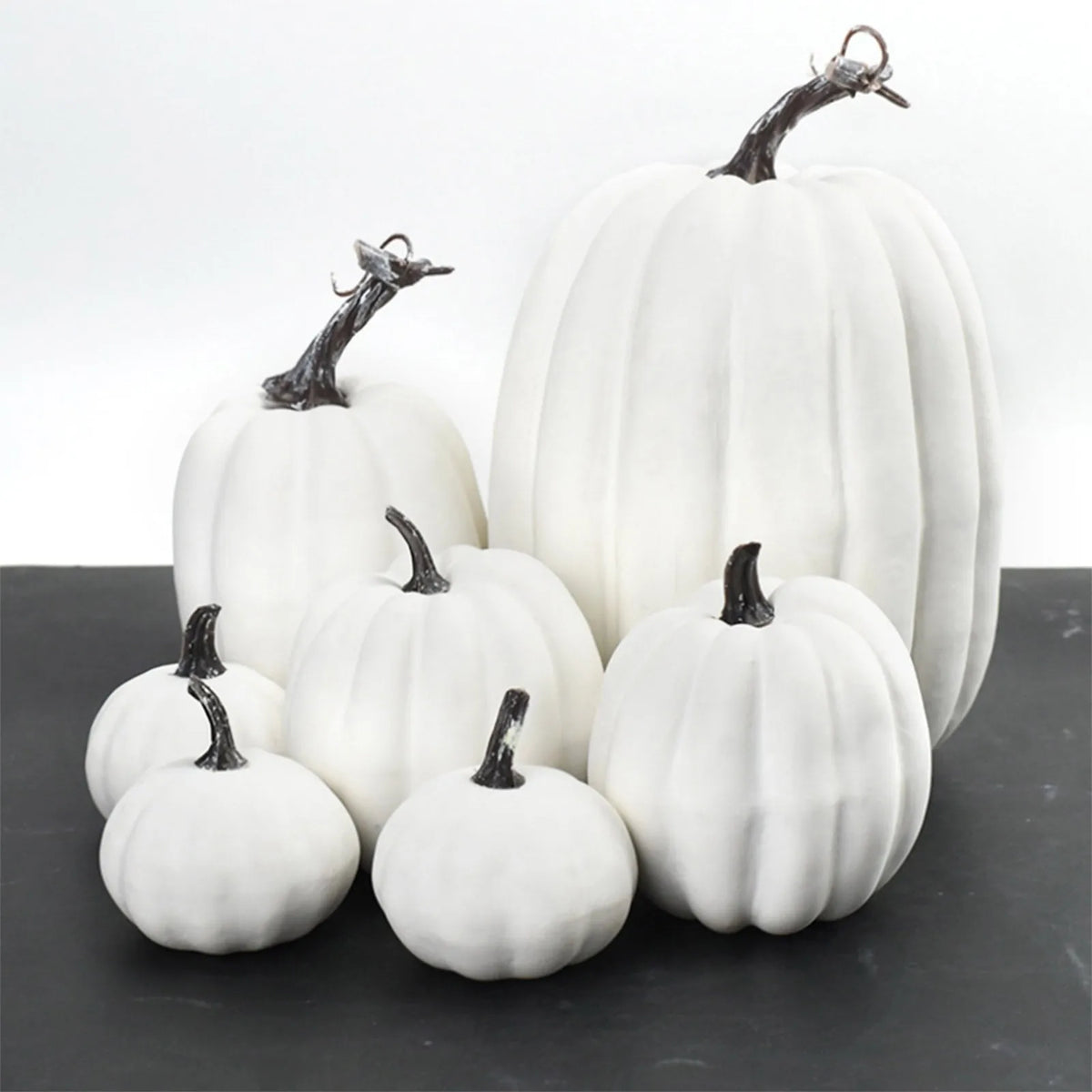 A set of seven white artificial pumpkins with dark stems arranged on a dark surface.