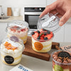 A hand holds a clear round dessert cup with a lid and spoon, filled with layered dessert, on a wooden board.